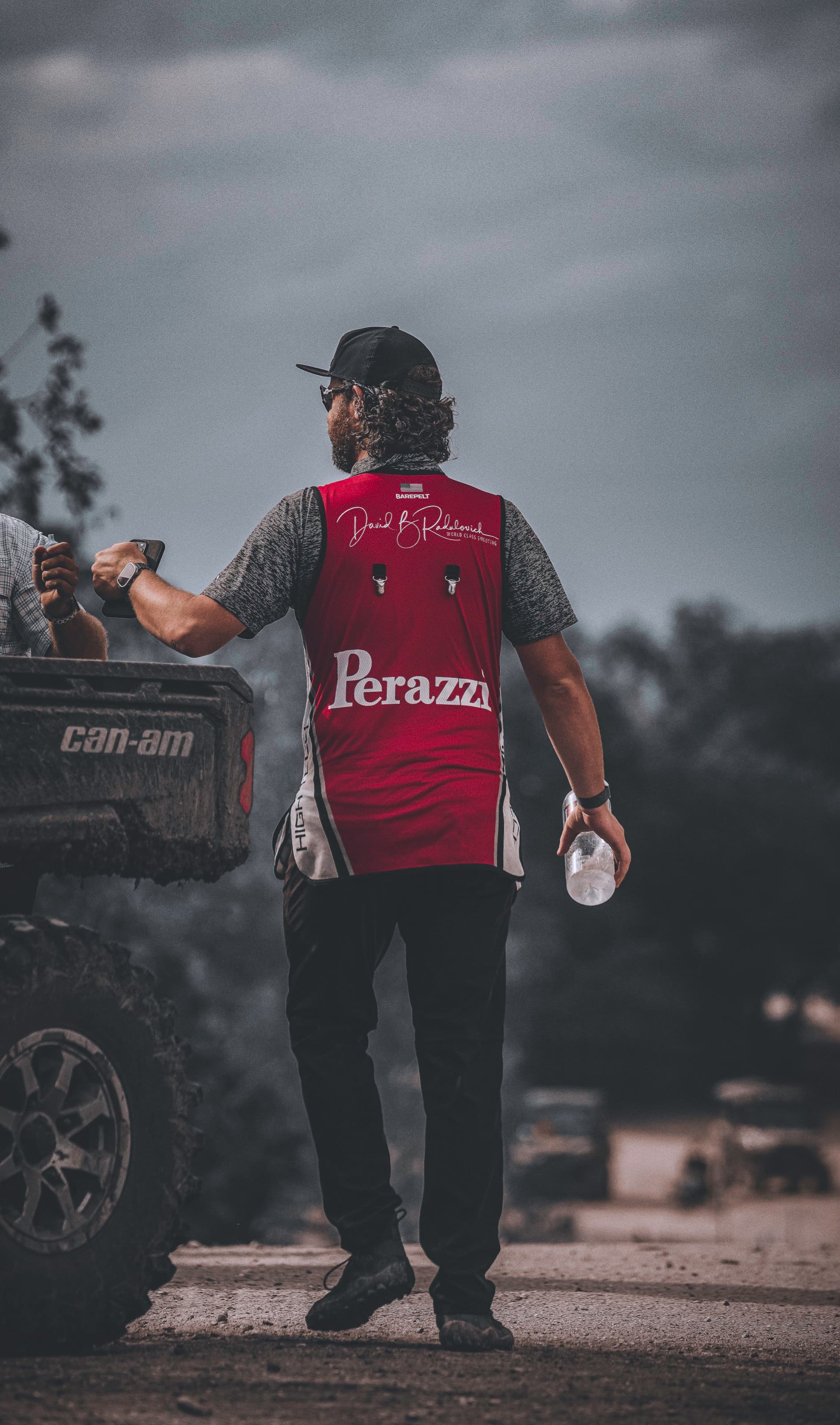 A shooter in a red vest walks away into a gray field with a water bottle in hand.