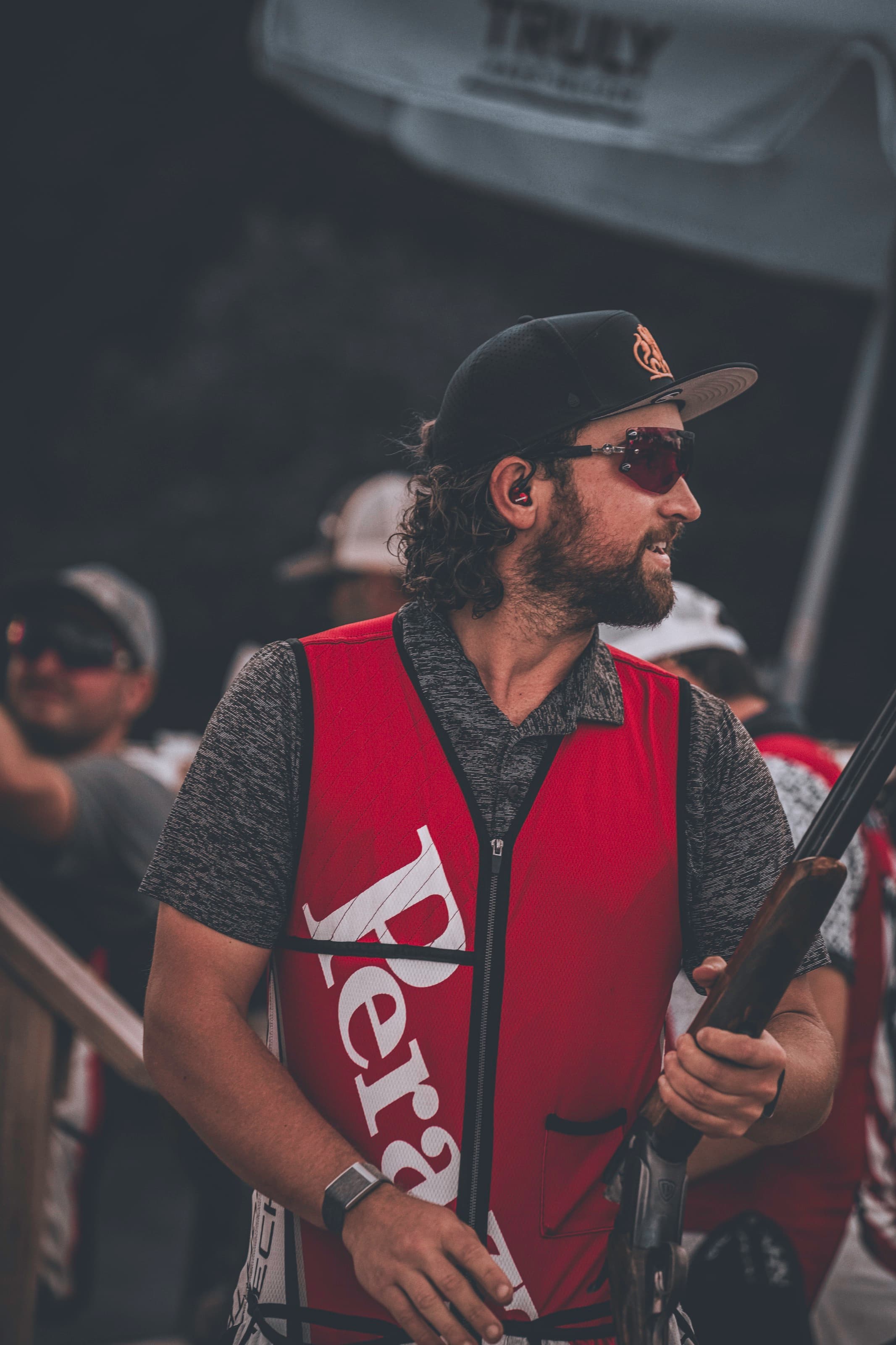 A shooter in a red vest holding a shotgun while looking toward the field.