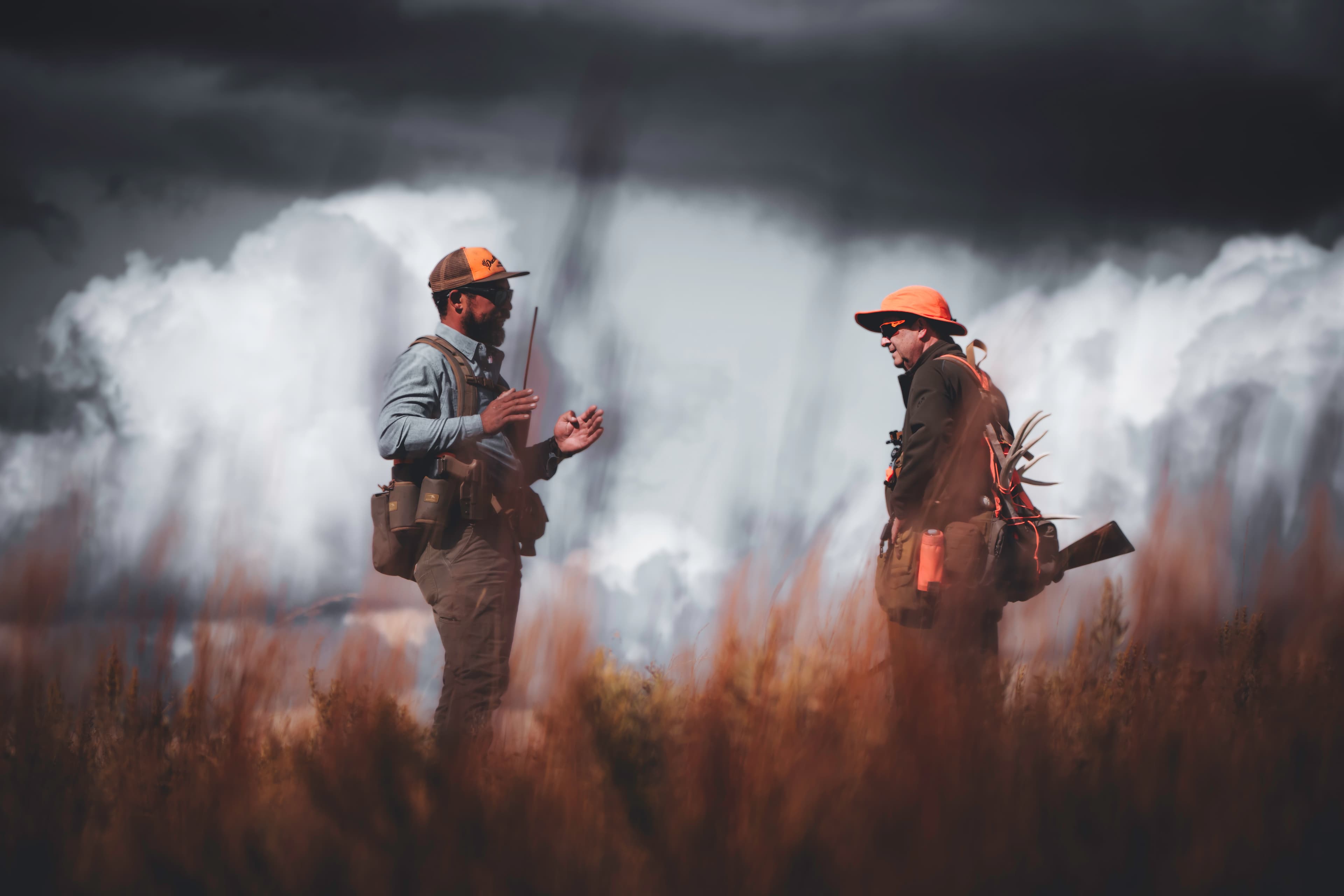 Two people in field gear speaking in front of a storm cloud.