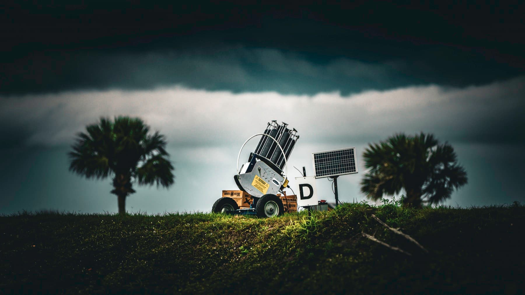 A clay target machine standing beneath a dark storm shelf in an open sporting field.