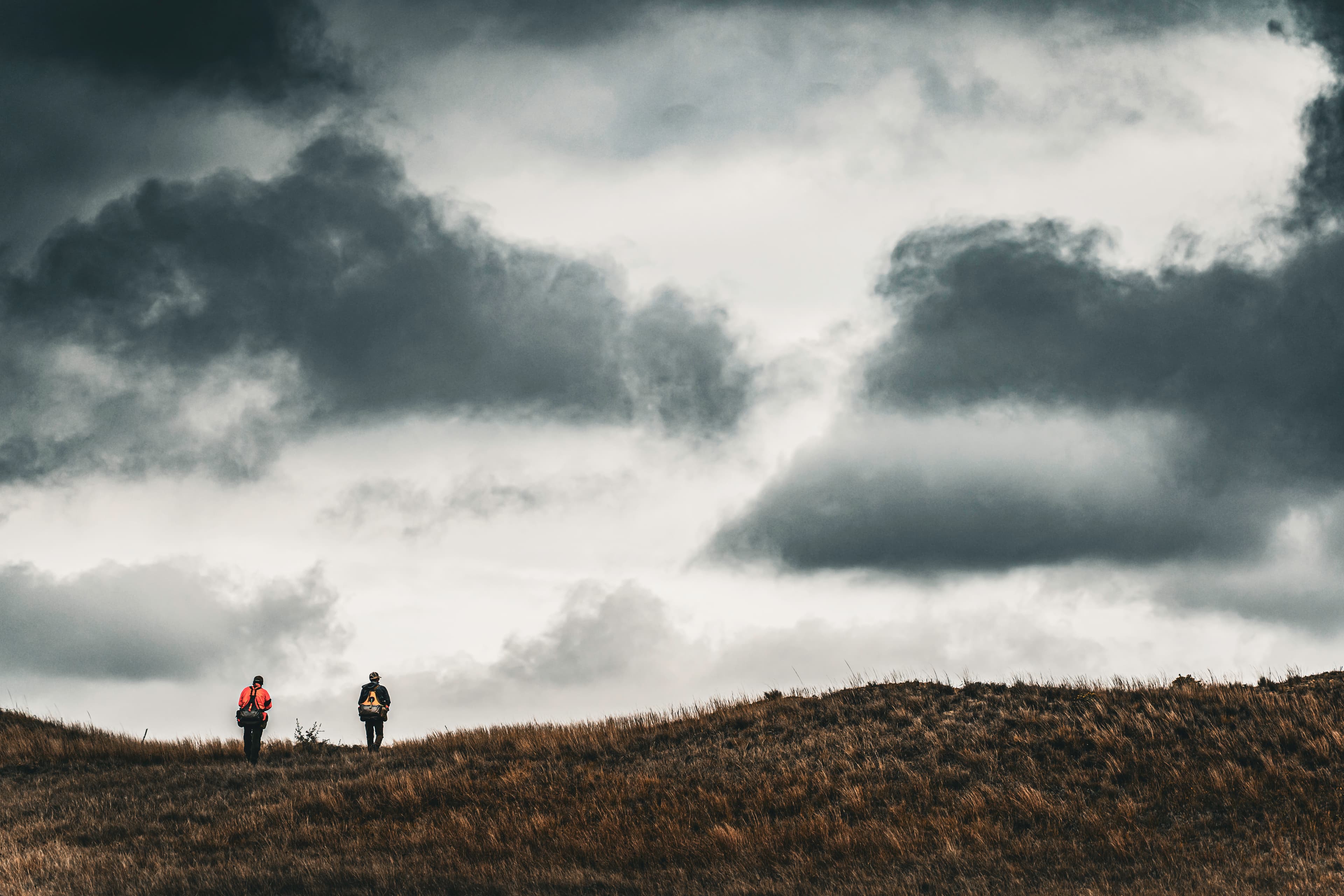 Two small figures walking across a windy ridge beneath a large cloud field.