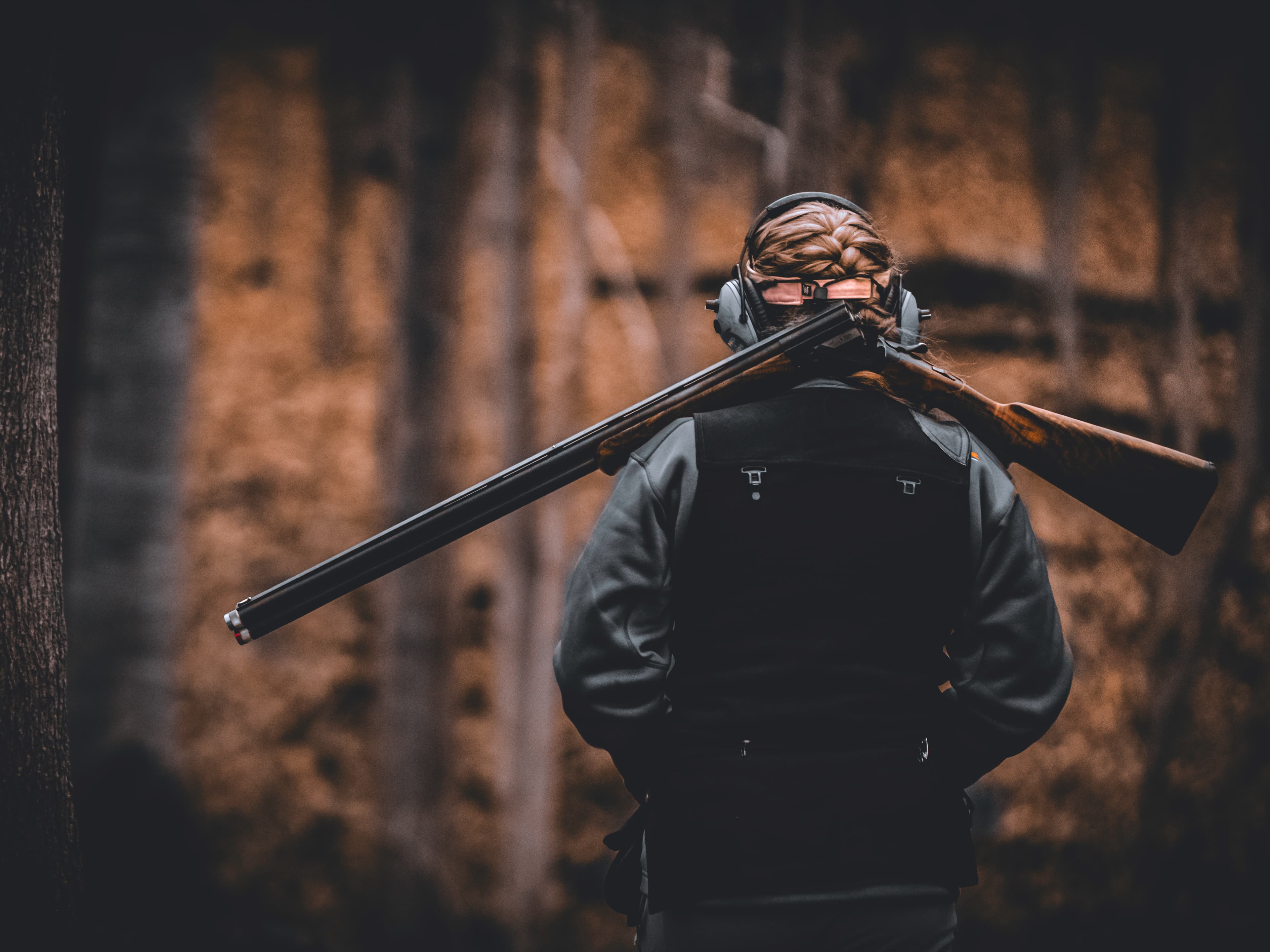 A person seen from behind in the woods with a shotgun resting across their shoulders.