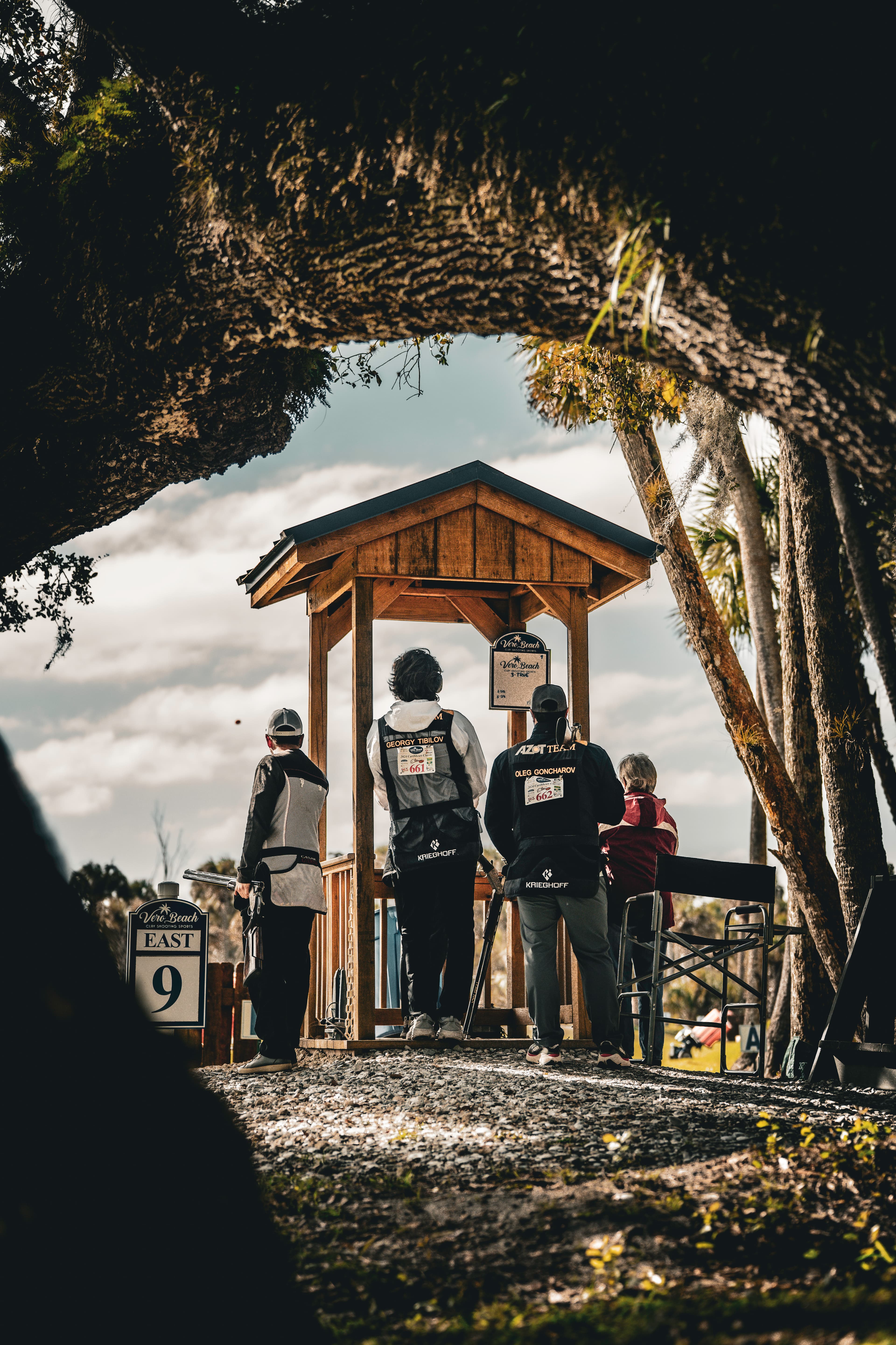 People standing near a shooting station beneath a dark tree canopy.